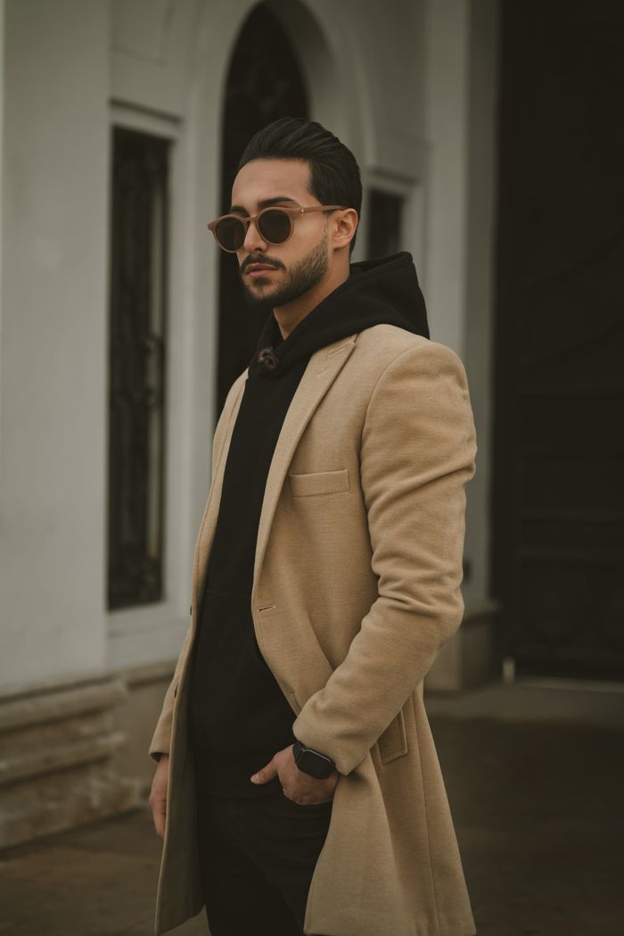 Man in beige coat posing outside an urban building, showcasing trendy fashion style.